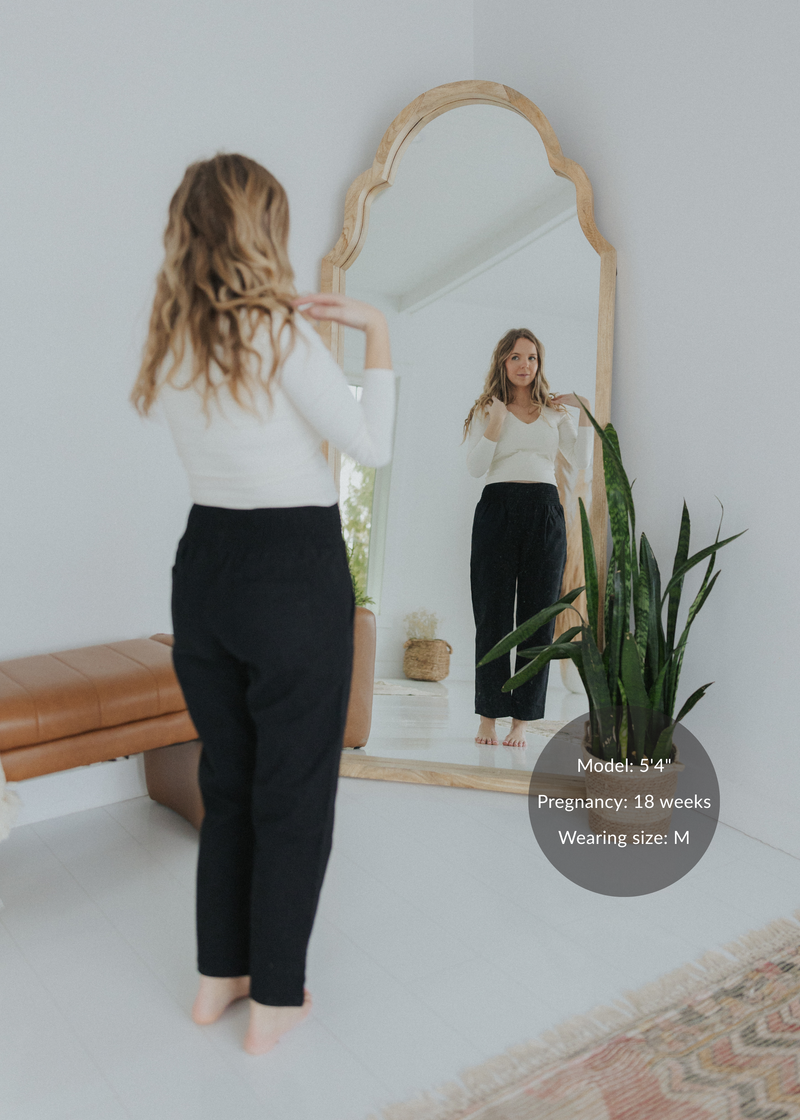 Woman in black smocked linen maternity pants and white top, reflected in a mirror.