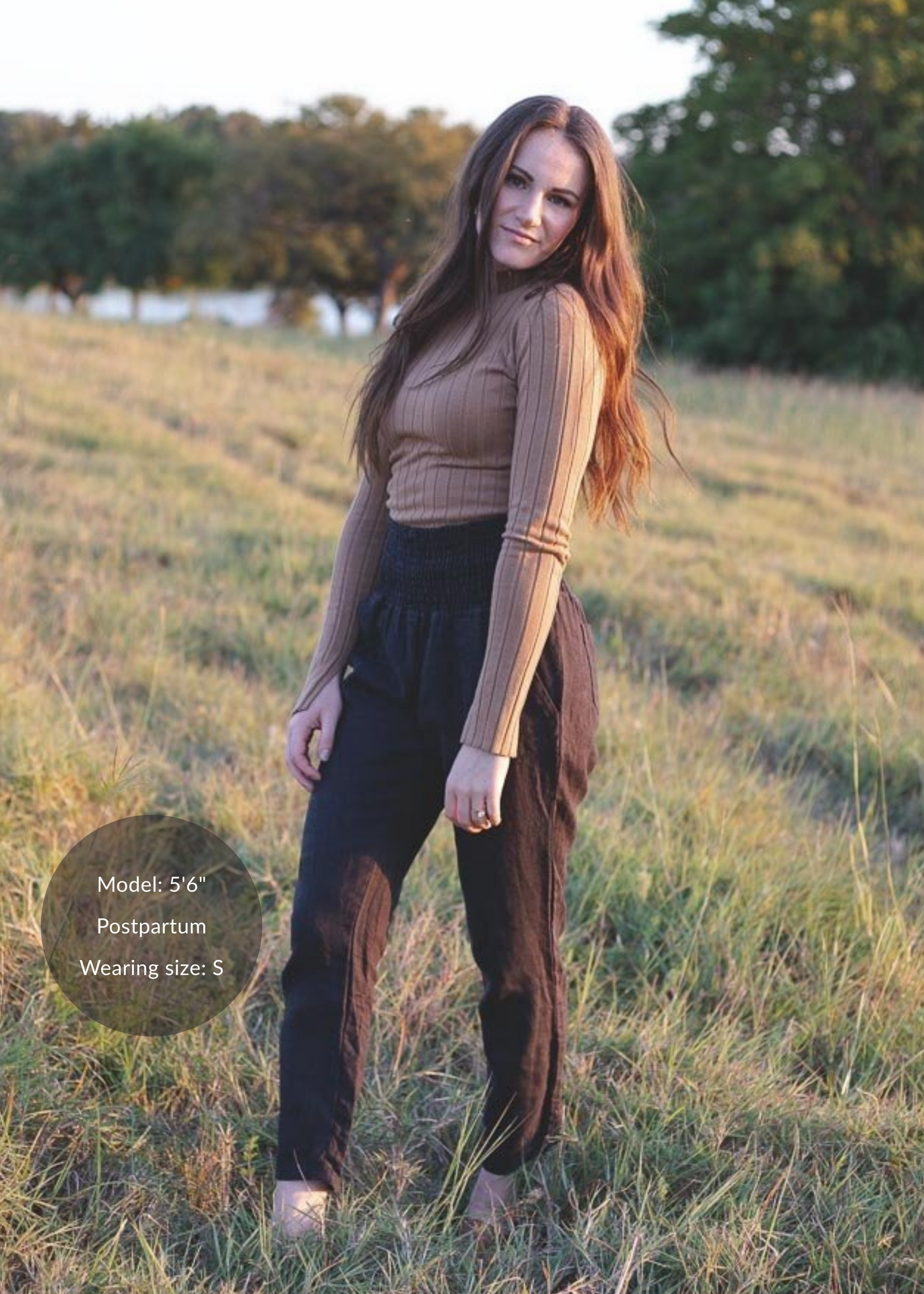 Woman in black smocked linen pants and a brown ribbed top standing in a grassy field.