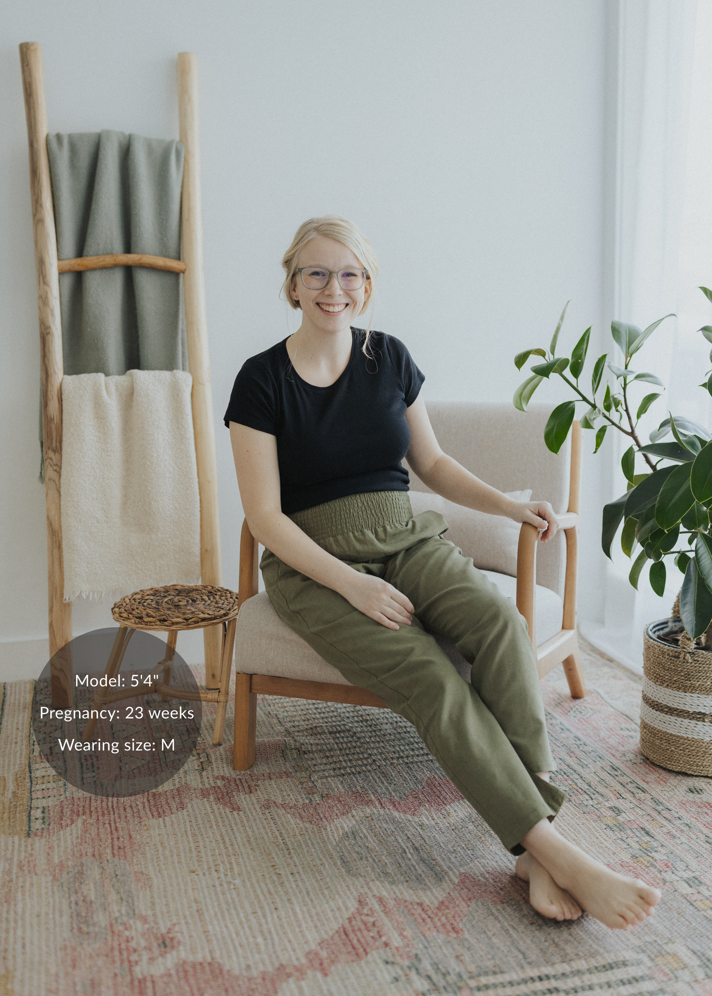 Smiling pregnant woman in olive green smocked linen pants and black tee, sitting in a chair.