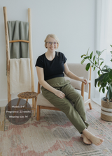 Smiling pregnant woman in olive green smocked linen pants and black tee, sitting in a chair.