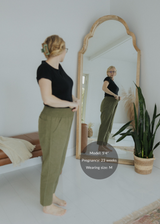 Pregnant woman in olive green smocked linen pants and black top, reflected in a mirror.