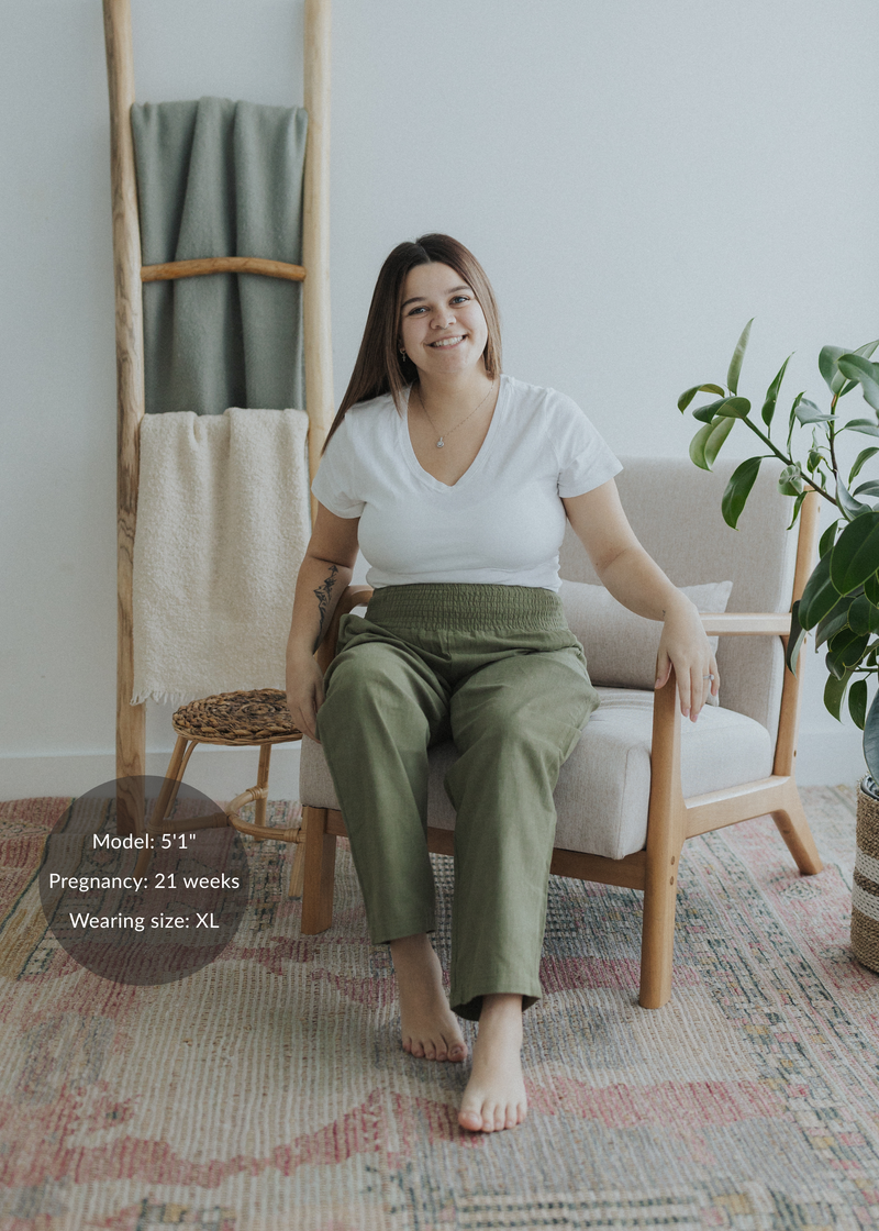 Smiling pregnant woman in olive green smocked linen maternity pants and white tee, seated.