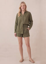 Smiling woman in olive green cotton gauze button-up shirt and matching smocked shorts.