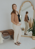 Smiling pregnant woman in natural linen smocked pants, black top, and brown cardigan.
