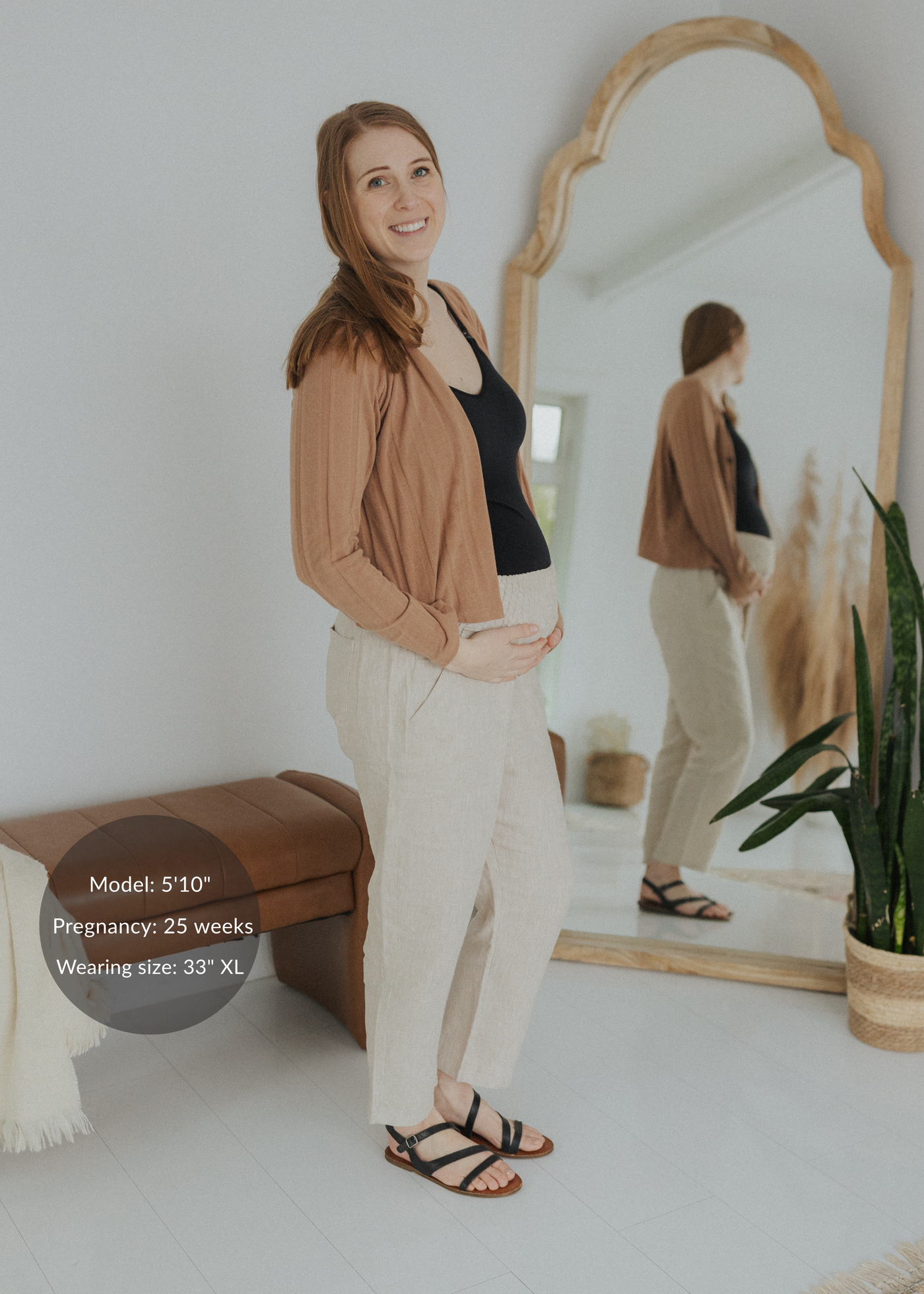 Smiling pregnant woman in natural linen smocked pants, black top, and brown cardigan.