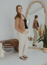 Smiling pregnant woman in natural linen smocked pants, black top, and brown cardigan.
