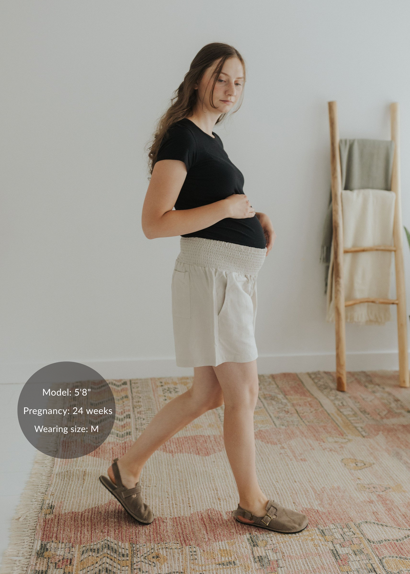 Pregnant model wearing light beige smocked cotton-linen shorts with deep pockets and a black t-shirt.