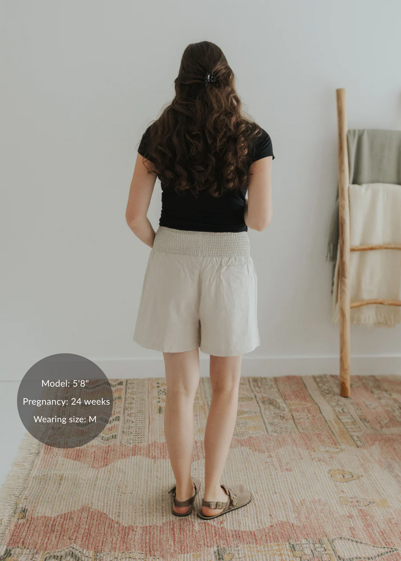 Back view of a pregnant model wearing light beige smocked cotton-linen shorts and a black top.