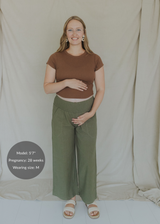 Smiling pregnant woman in olive wide-leg linen pants with a smocked waistband and a brown top.