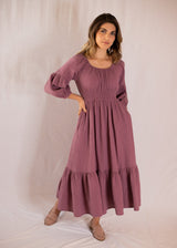 Woman in Eliza Maternity and Nursing Dress, a mauve cotton gauze maxi dress with smocked waist, balloon sleeves, and ruffle skirt.