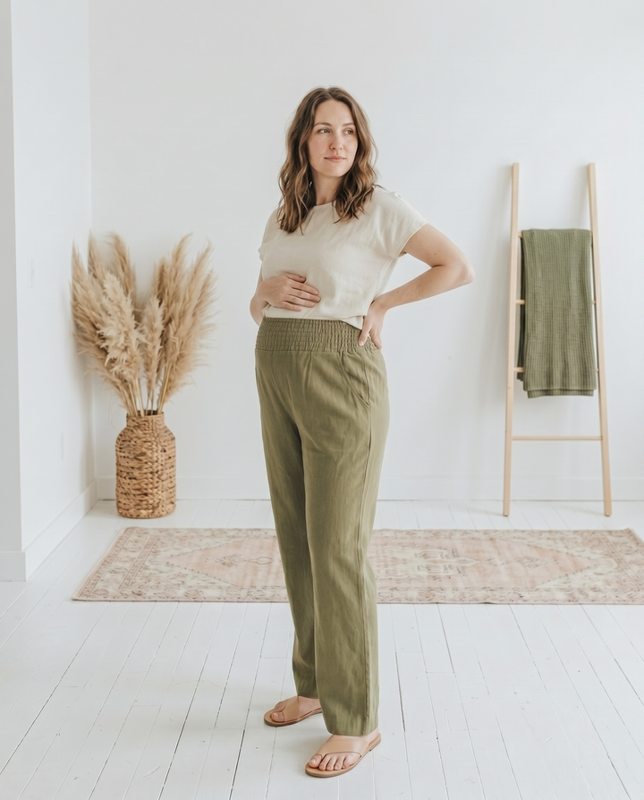 Pregnant model in olive green straight-leg smocked linen maternity pants with a smocked waistband and side pockets, standing in a bright white loft interior.