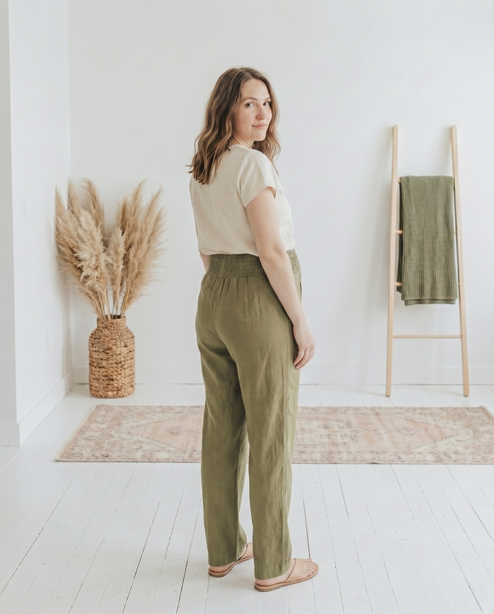Back view of pregnant model in olive green straight-leg smocked linen maternity pants, showing the smocked waistband, in a white loft interior.
